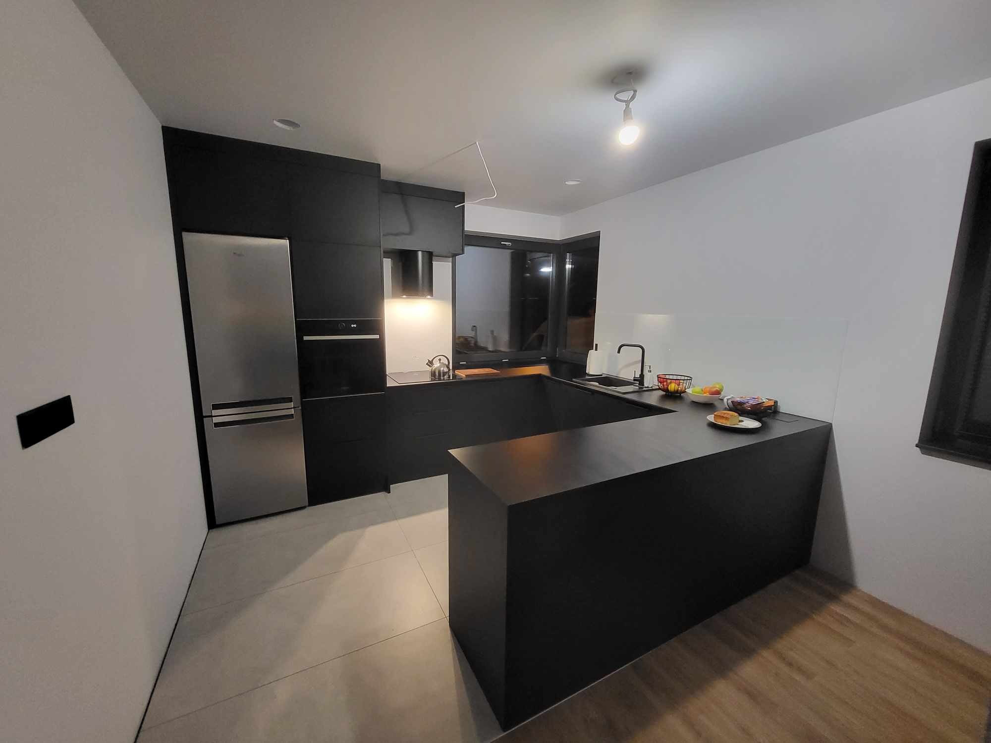 Nowoczesna, czarna kuchnia z wyspą i srebrną lodówką. Minimalistyczny design, jasne ściany, drewniana podłoga w tle. Oświetlenie punktowe i naturalne światło z okna.