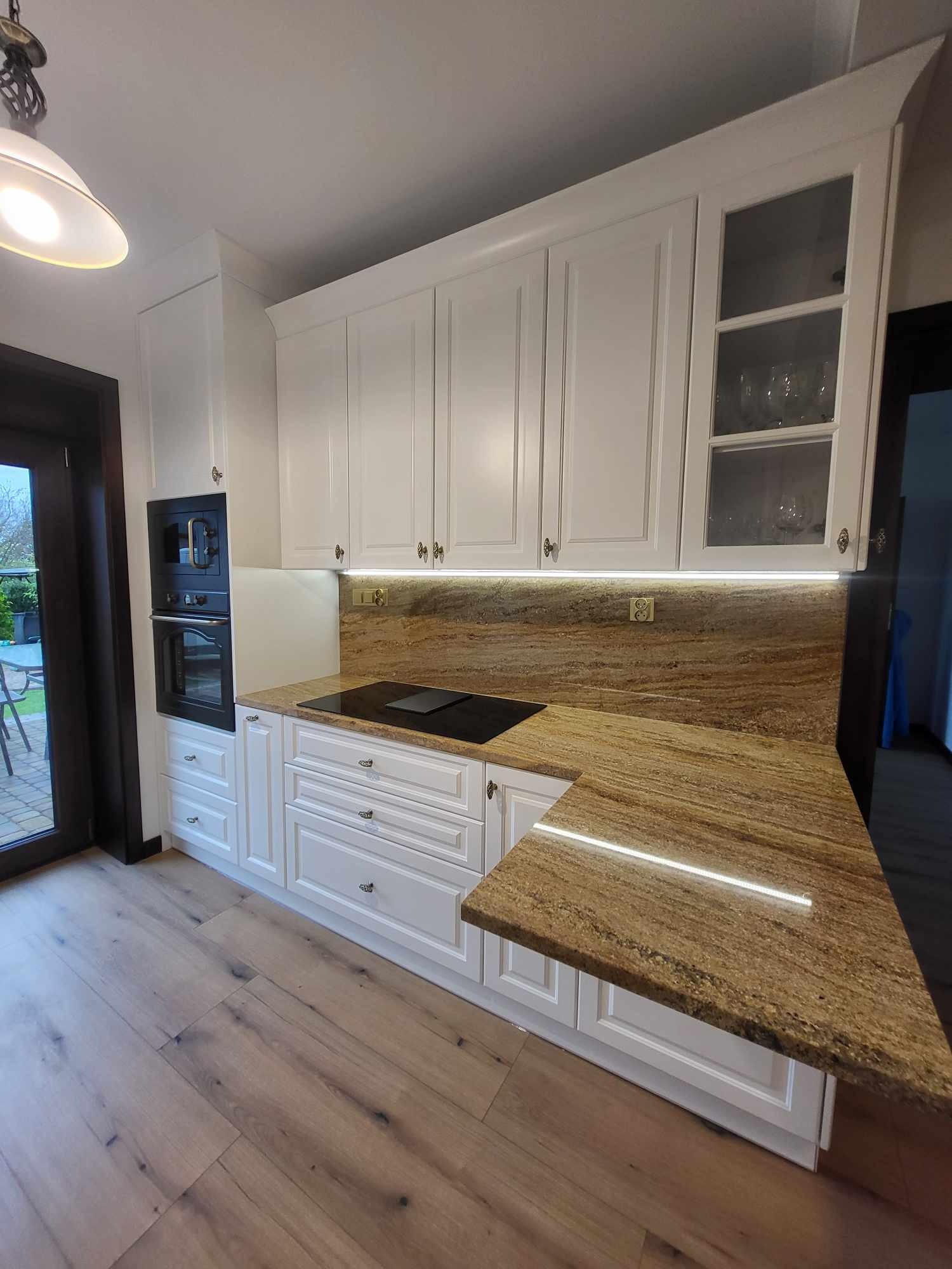 Biała kuchnia z drewnianym blatem i oświetleniem LED. Widoczne szafki wiszące z przeszklonymi frontami, piekarnik w zabudowie i drewniana podłoga.