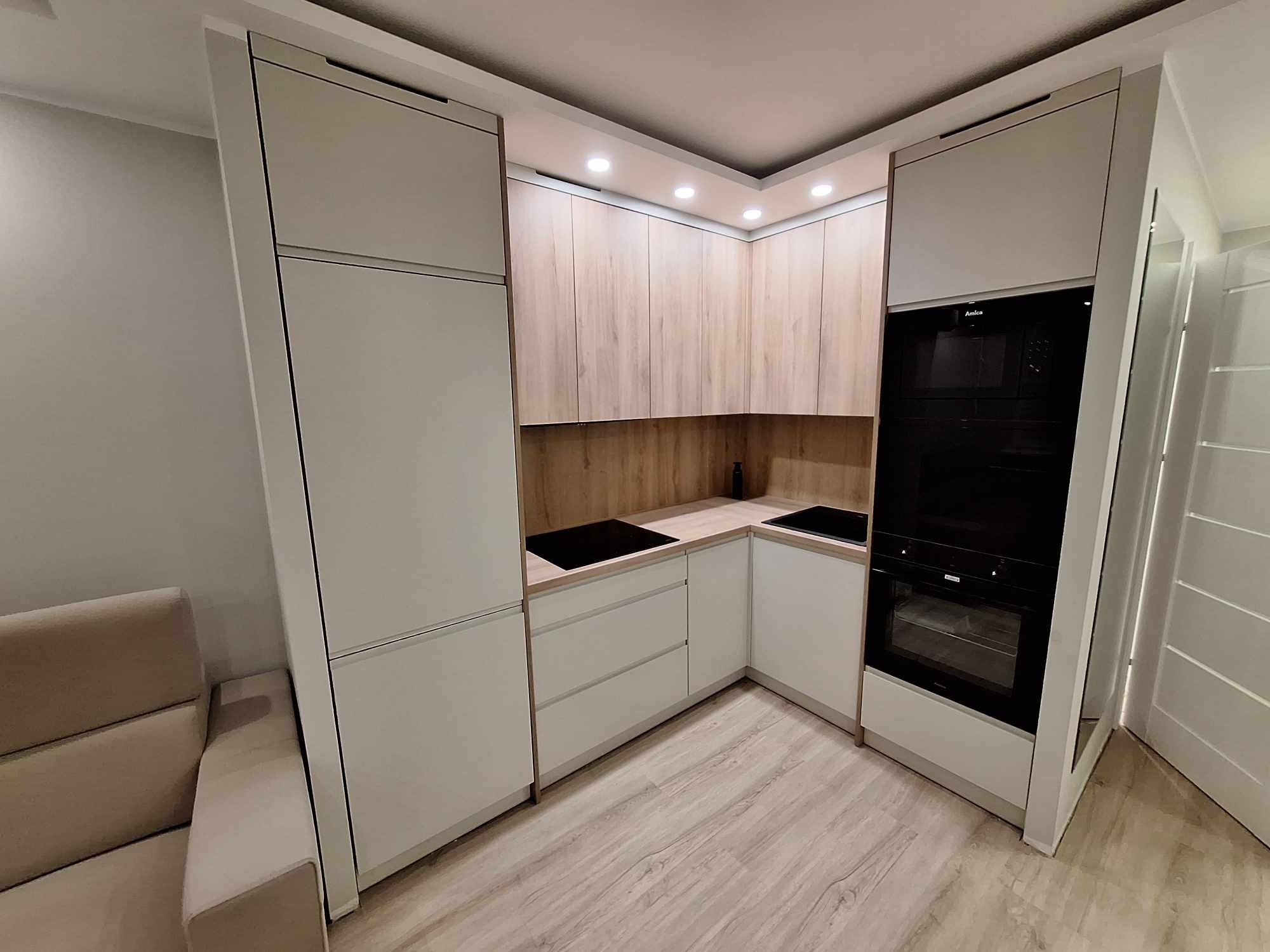 Jasna kuchnia w zabudowie z drewnianymi akcentami, oświetleniem punktowym i sprzętem AGD marki Amica. Widok na minimalistyczne fronty i drewnopodobne wykończenie.