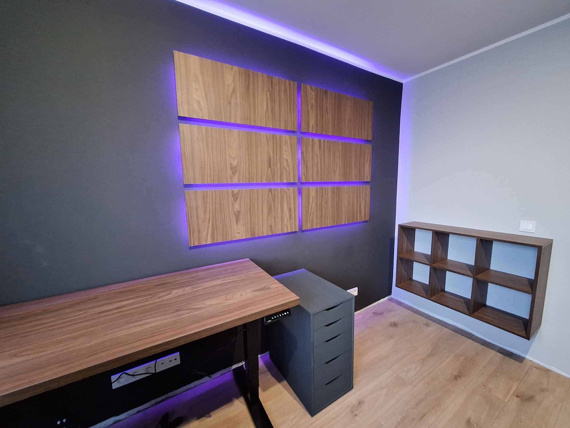 Drewniany blat biurka z regulacją wysokości, szara komoda z szufladami, modułowa półka i dekoracyjne panele ścienne z podświetleniem LED w nowoczesnym gabinecie.