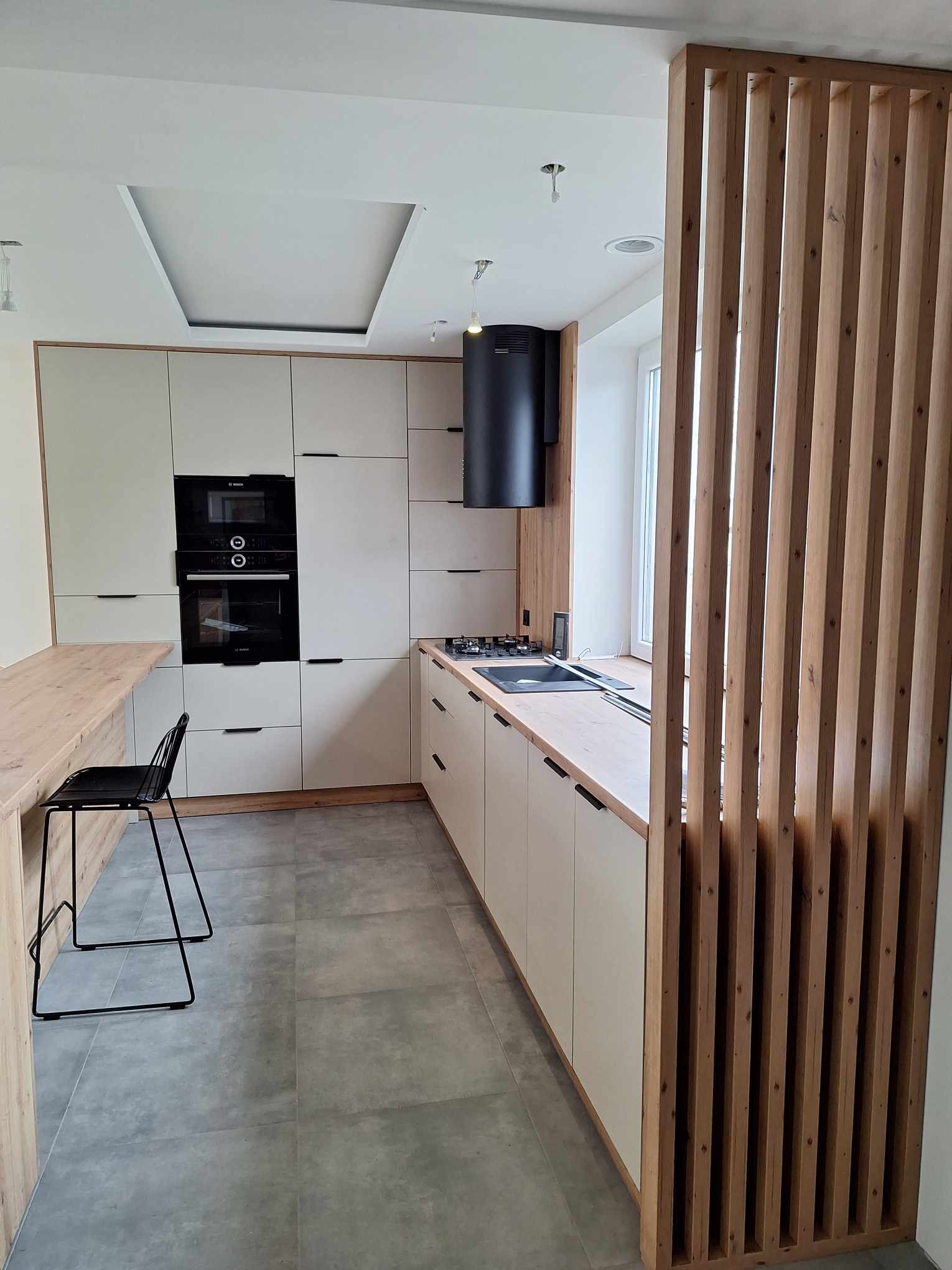 Nowoczesna kuchnia z zabudową w odcieniach beżu i drewna, z czarnymi akcentami. Widoczna drewniana ścianka działowa. Realizacja w Wanatach.