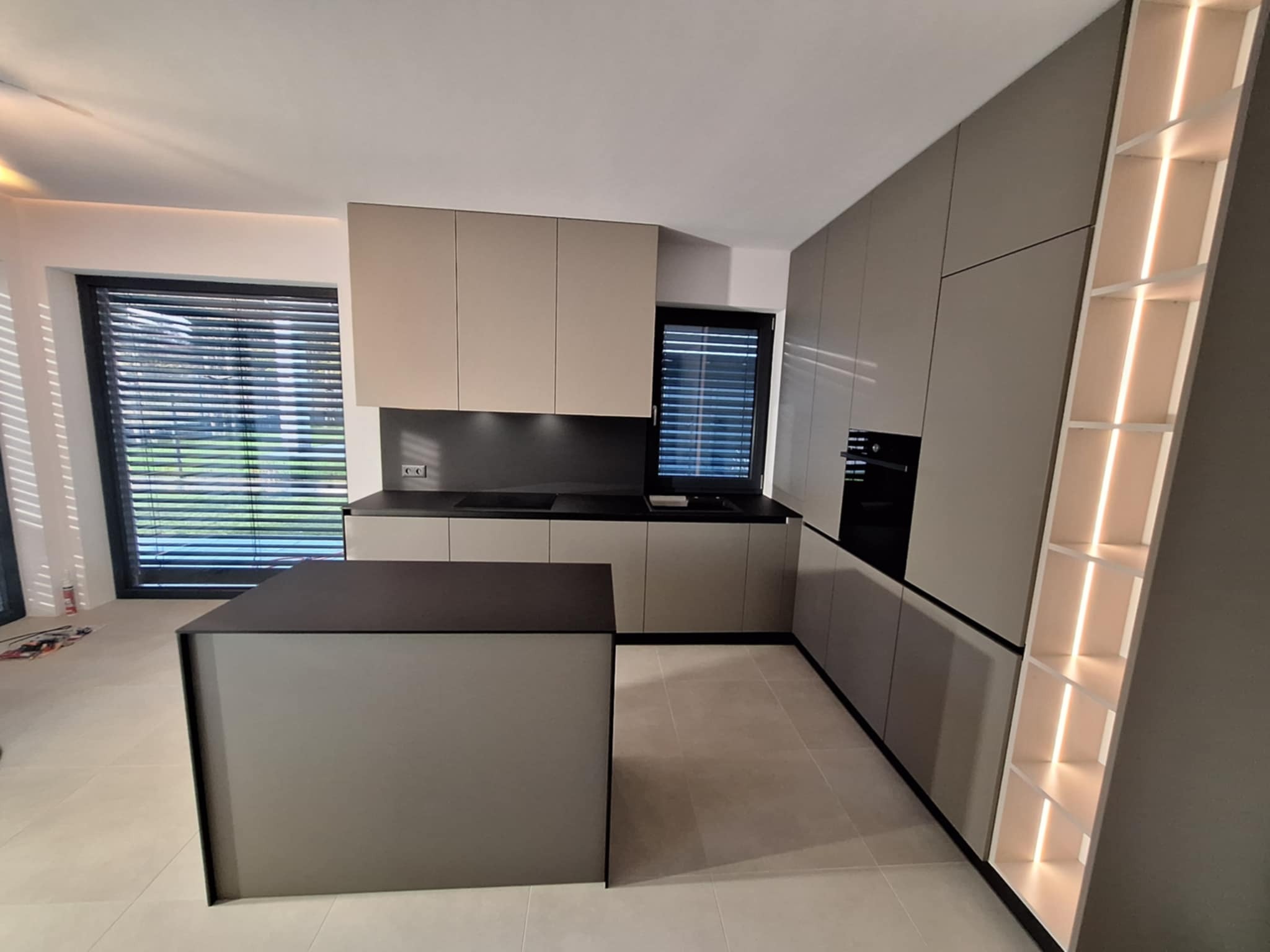 Nowoczesna kuchnia z wyspą w odcieniach szarości i beżu. Minimalistyczne fronty, czarny blat, oświetlenie LED w regale. Duże okno z żaluzjami w tle.