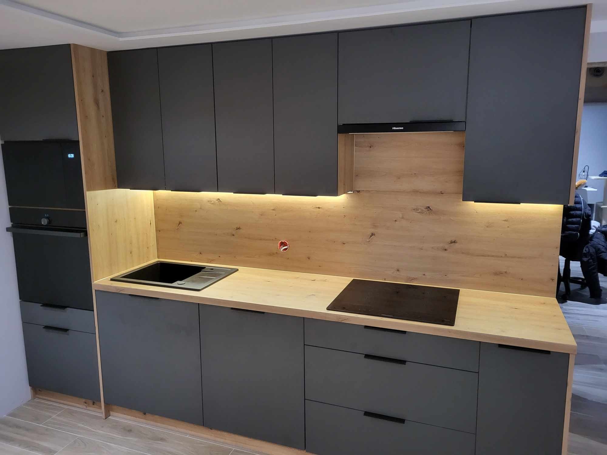 Nowoczesna kuchnia z szarymi frontami i drewnianym blatem. Oświetlenie LED podkreśla fakturę drewna. Widoczny zlew, płyta indukcyjna i piekarnik.