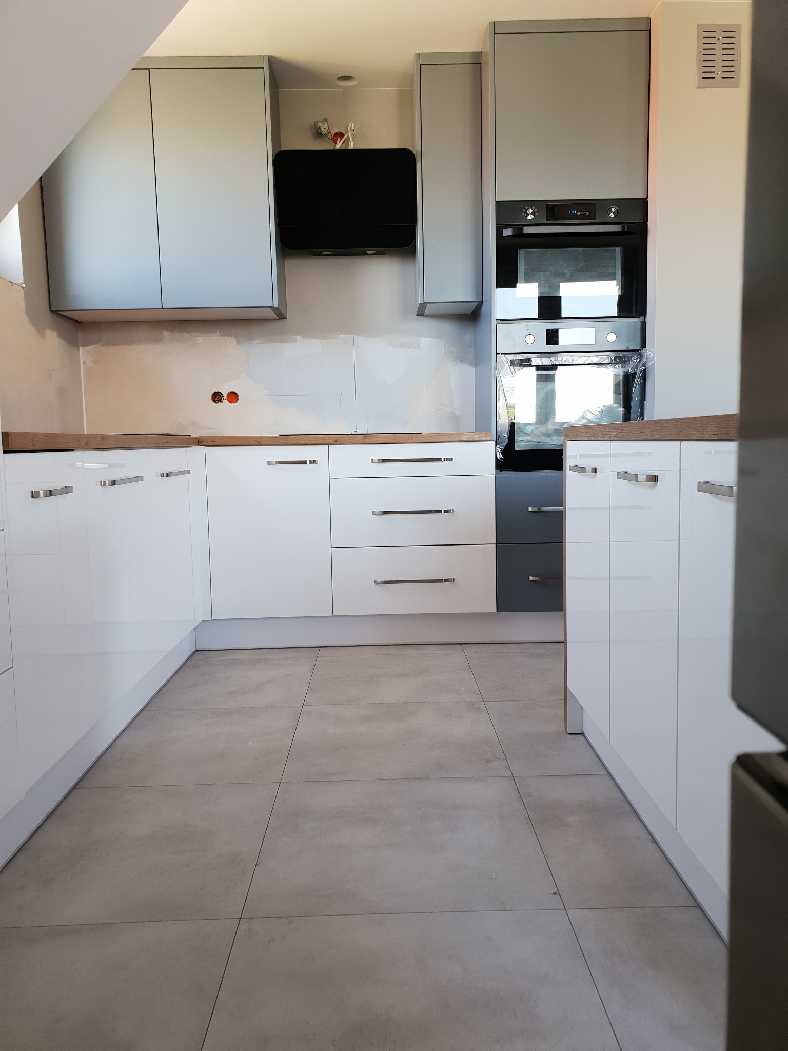 Nowoczesna kuchnia z białymi, lakierowanymi frontami i szarymi szafkami wiszącymi. Widoczny okap i piekarnik w zabudowie. Blat drewnopodobny. Częściowo wykończone ściany.