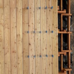 Sanadom Marcin Linowiecki - Montaż desek tarasowych z modrzewia syberyjskiego na drewnianej konstrukcji nośnej, widoczne niebieskie klipsy montażowe.