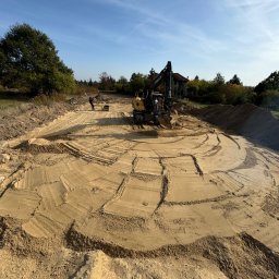 Sanadom Marcin Linowiecki - Przygotowanie terenu pod budowę z użyciem koparki i zagęszczarki gruntu, widoczne ślady gąsienic i równomiernie ubita warstwa piasku.