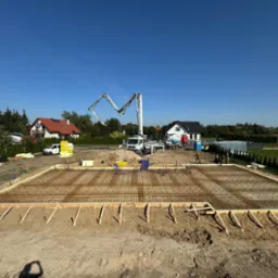 Zbrojenie przygotowane do wylania fundament&oacute;w pod budowę domu jednorodzinnego, widoczna pompa do betonu i ekipa budowlana w tle.