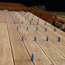 Sanadom Marcin Linowiecki - Układanie desek tarasowych z użyciem niebieskich dystansów, widoczna konstrukcja nośna tarasu i fragment ogrodzenia w tle.