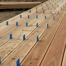 Sanadom Marcin Linowiecki - Deski tarasowe z niebieskimi elementami dystansowymi, ułożone równolegle, widoczne przygotowania do montażu, w tle hałda piasku.