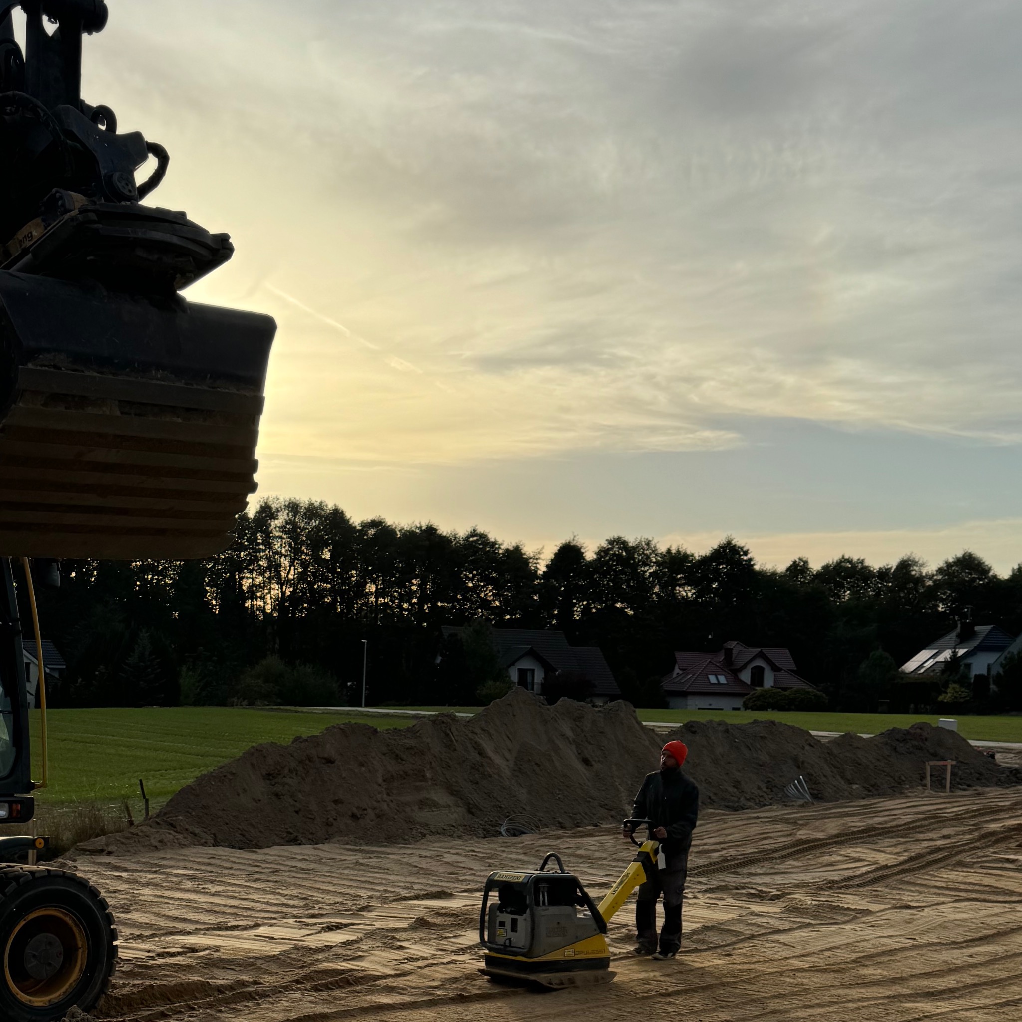 Operator w kasku zagęszcza piasek zagęszczarką wibracyjną na placu budowy, w tle widoczny sprzęt budowlany i hałda piasku, a dalej domy i drzewa pod wieczornym niebem.