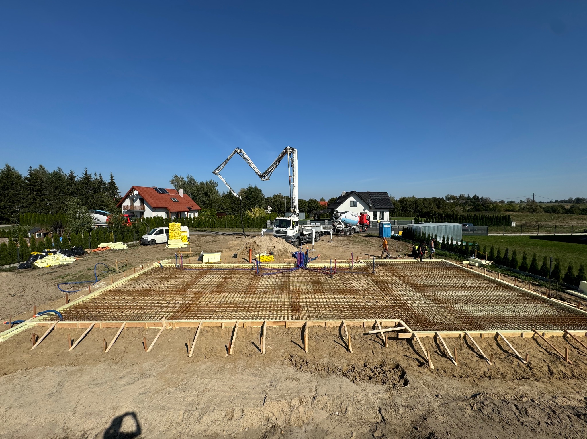 Zbrojenie przygotowane do wylania fundamentów pod budowę domu jednorodzinnego, widoczna pompa do betonu i ekipa budowlana w tle.