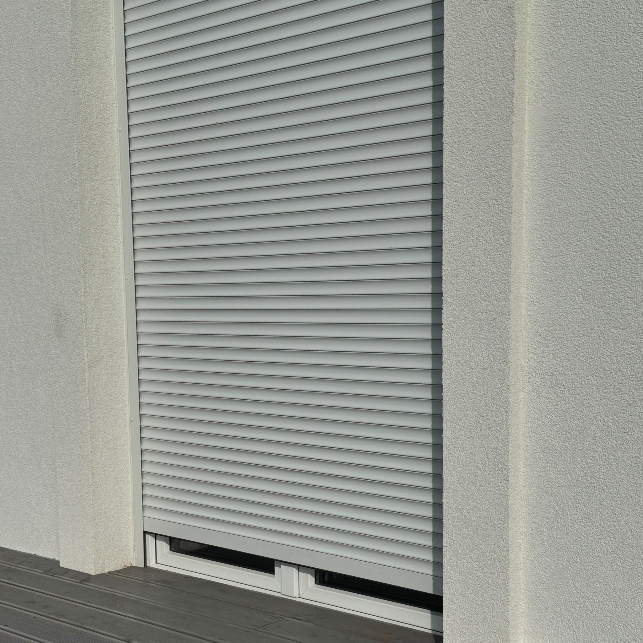 Zamknięta, zewnętrzna roleta okienna w kolorze szarym, zamontowana w elewacji budynku o jasnej, ziarnistej strukturze, widoczny fragment szarego tarasu.