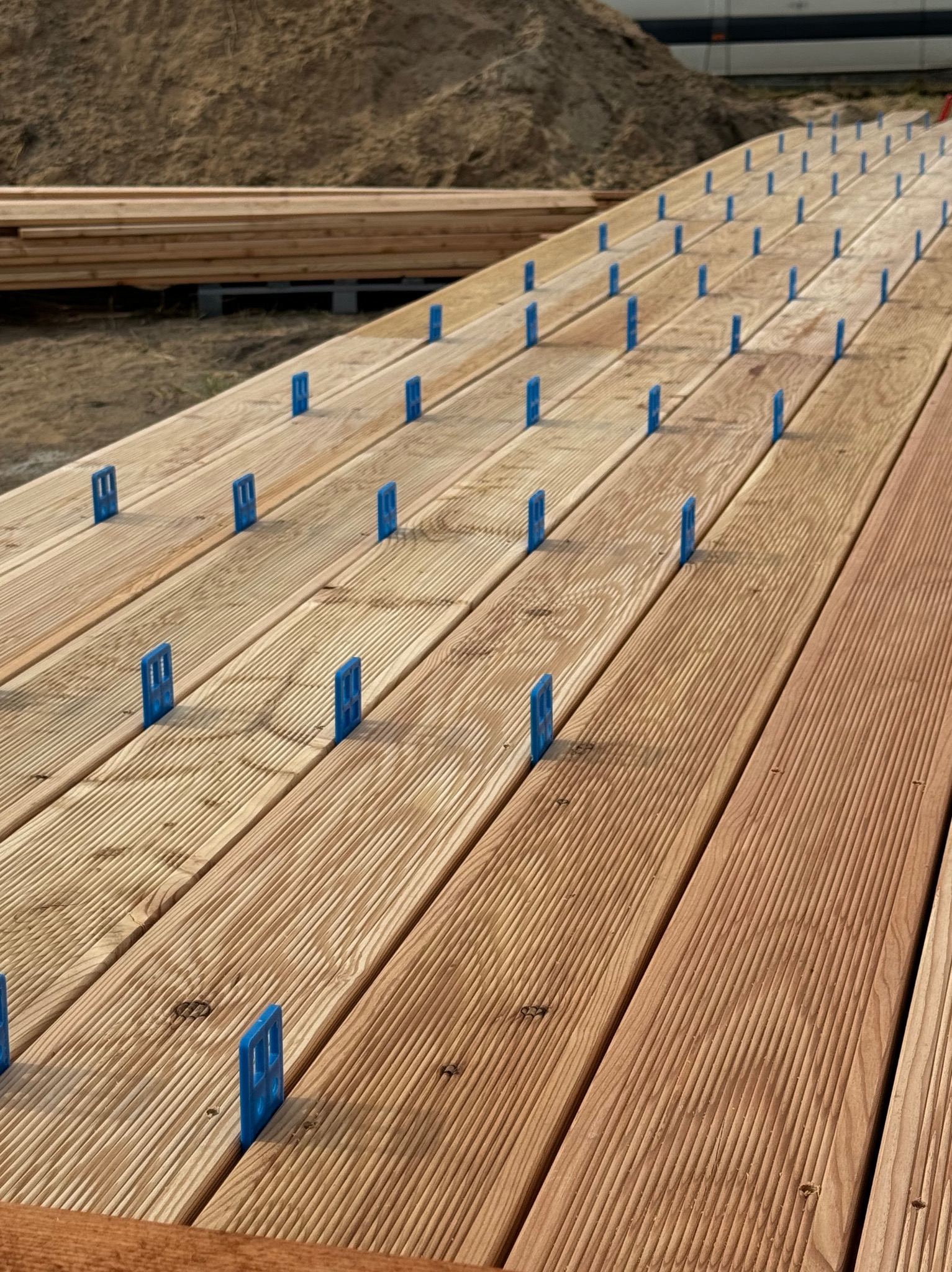 Deski tarasowe z niebieskimi elementami dystansowymi, ułożone równolegle, widoczne przygotowania do montażu, w tle hałda piasku.
