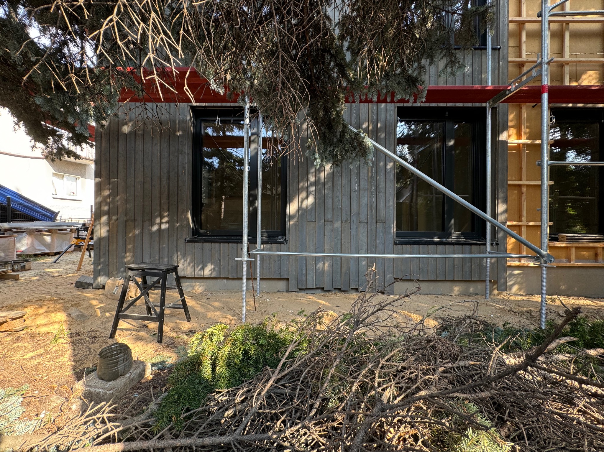 Budynek w trakcie rozbudowy z drewnianą elewacją, widoczne rusztowania, okna z czarnymi ramami, sterta gałęzi na pierwszym planie i drabinka.