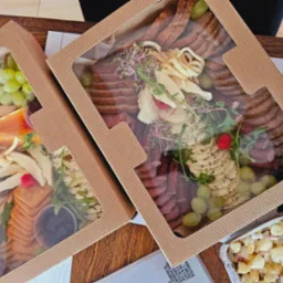 Dwa pudełka z przekąskami: w jednym owoce i sery, w drugim wędliny i sery, udekorowane zieleniną, obok sałatka ziemniaczana w aluminiowym pojemniku.