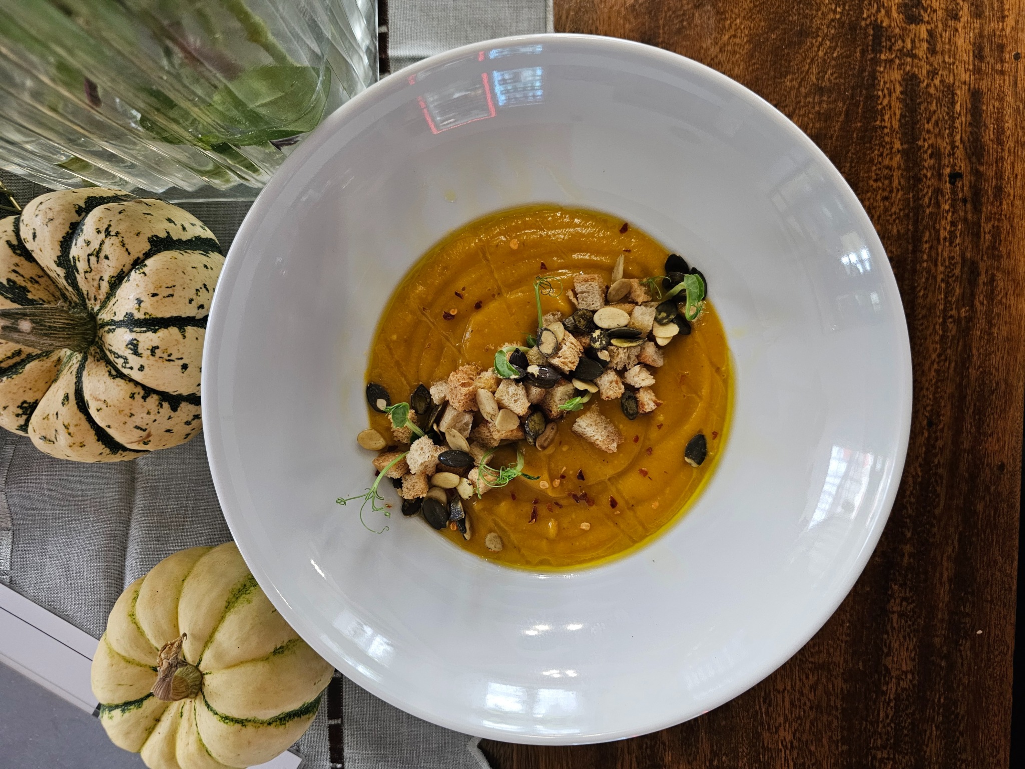 Krem z dyni w białym talerzu, udekorowany grzankami, pestkami dyni i mikro-ziołami, obok dwie dekoracyjne dynie na szarym obrusie, widok z góry.