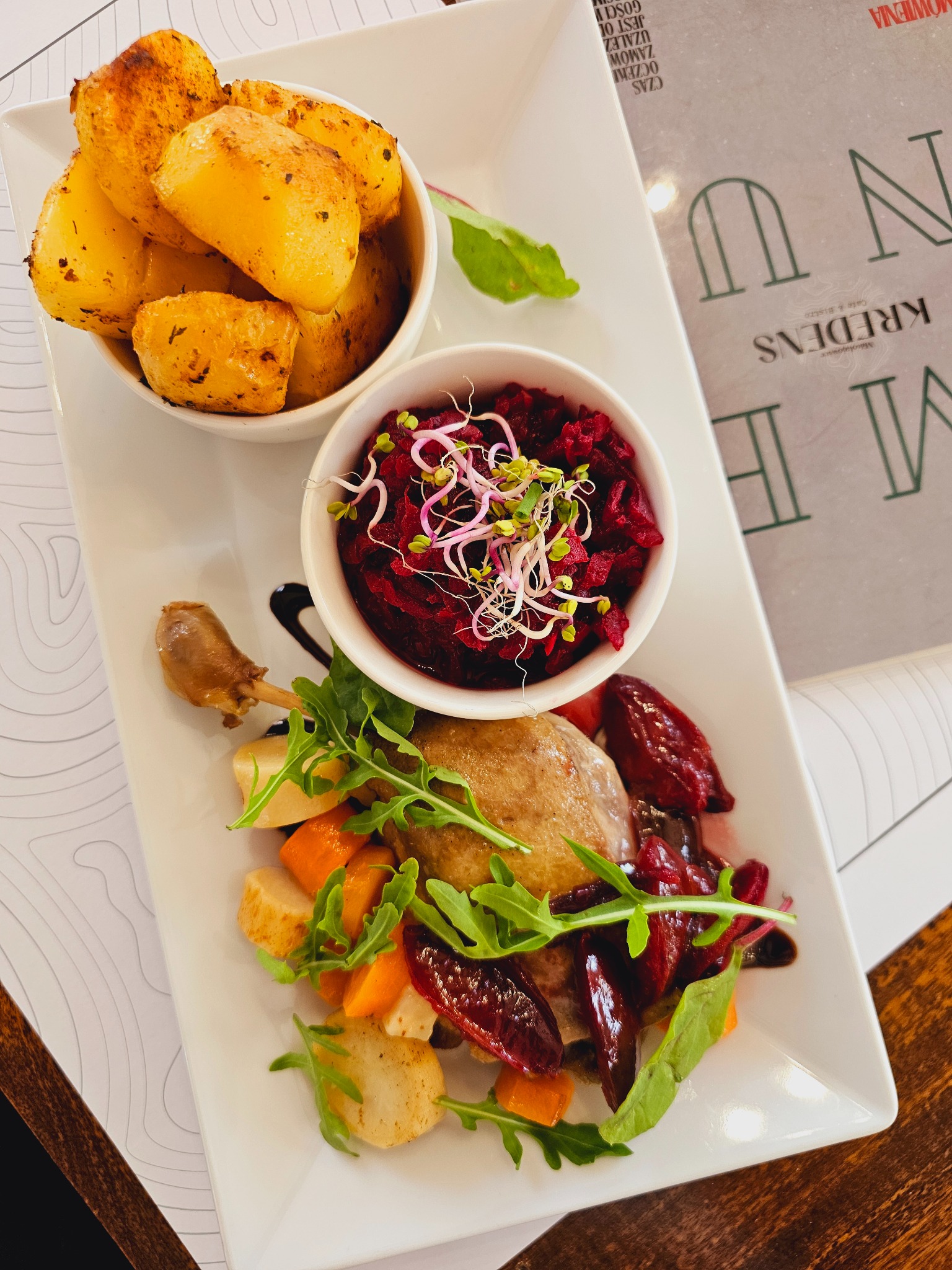 Elegancko podane danie z pieczoną kaczką, buraczkami z kiełkami, pieczonymi ziemniakami i warzywami na białym talerzu, obok menu restauracji.