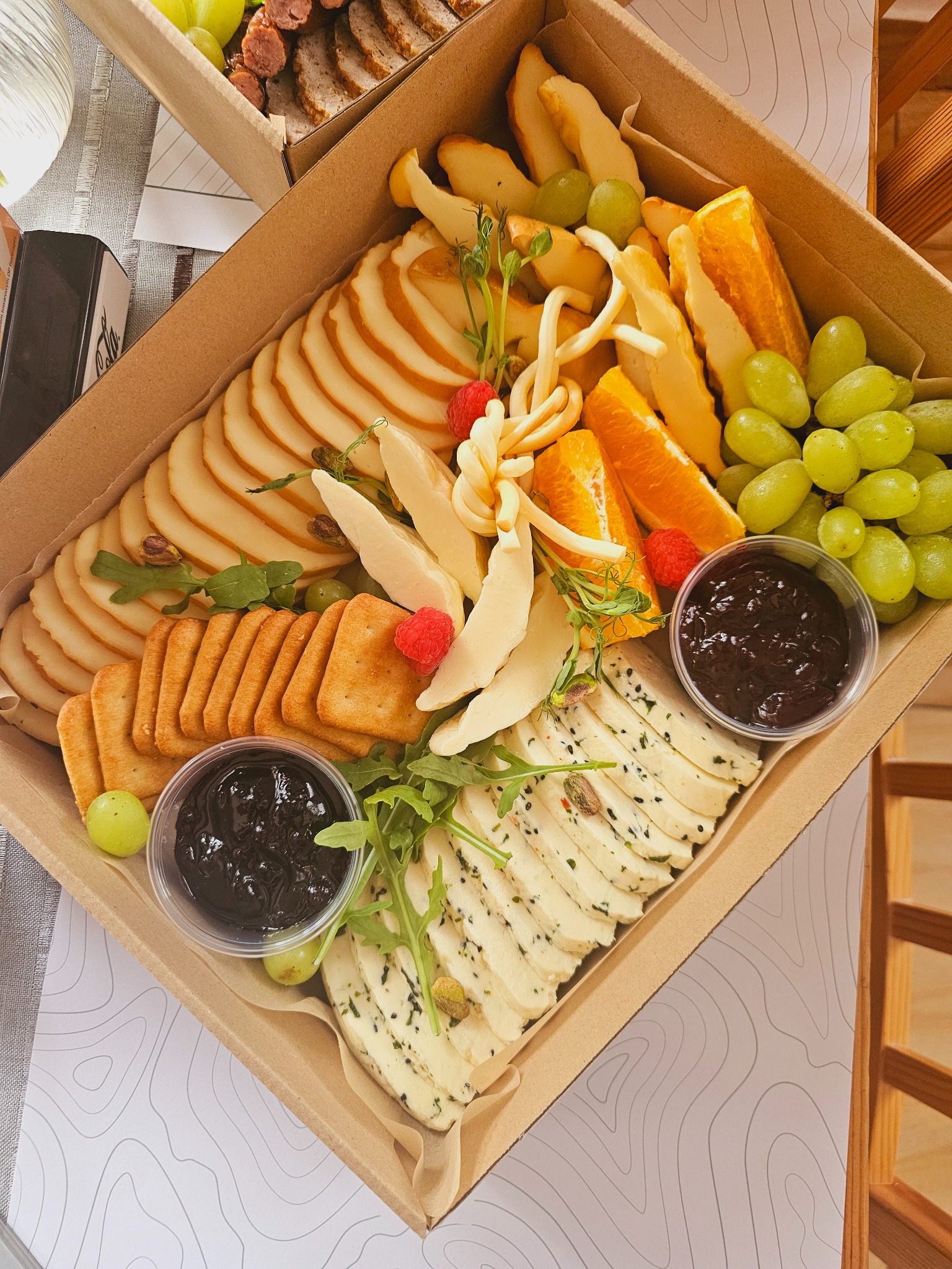 Elegancka kartonowa taca z selekcją serów (wędzony, pleśniowy, włoski), krakersów, winogron, pomarańczy, malin i konfitury, udekorowana świeżymi ziołami.