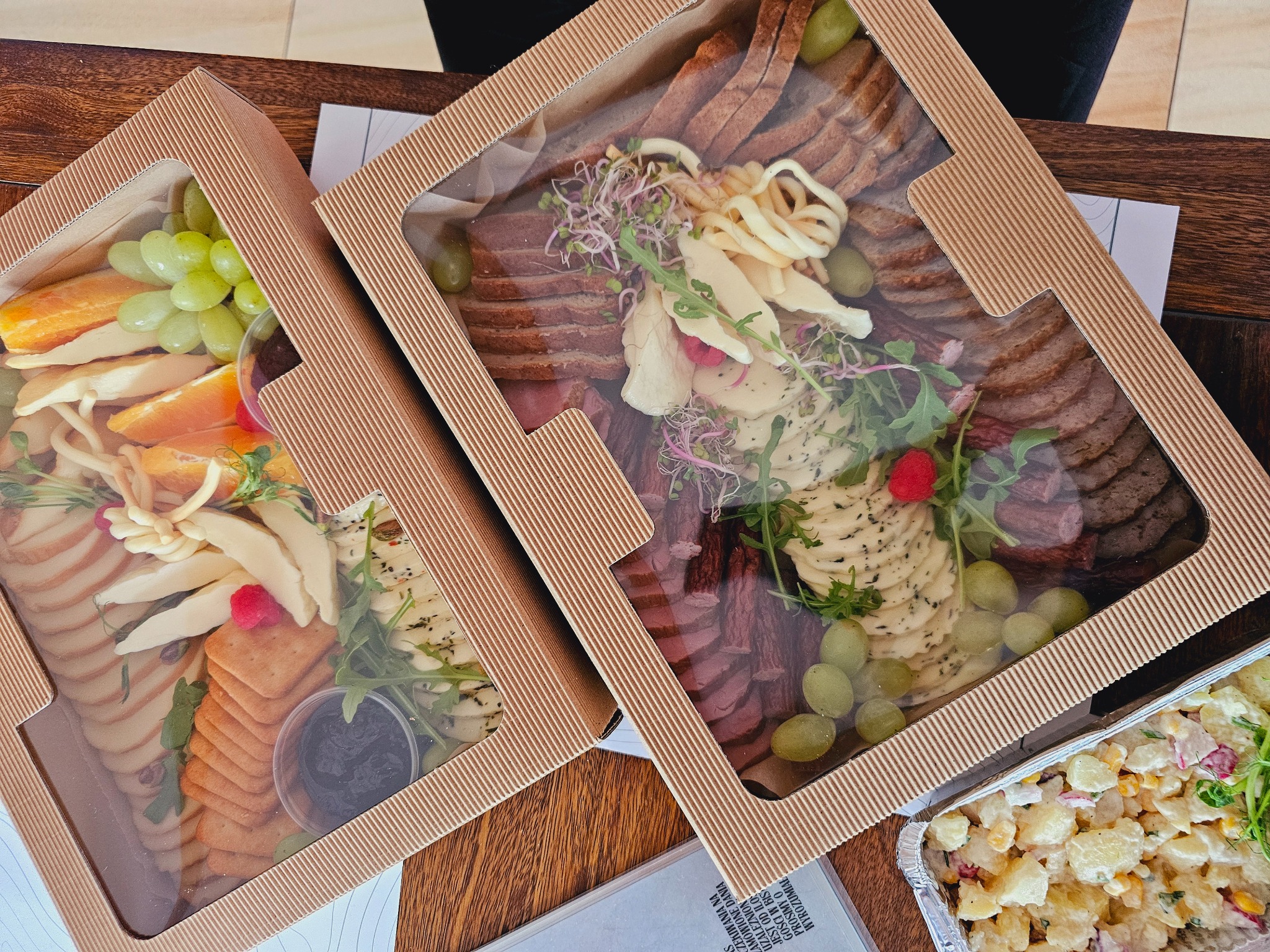 Dwa pudełka z przekąskami: w jednym owoce i sery, w drugim wędliny i sery, udekorowane zieleniną, obok sałatka ziemniaczana w aluminiowym pojemniku.