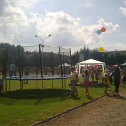 Dzieci skaczą na trampolinie z siatką zabezpieczającą podczas imprezy plenerowej z namiotami i balonami w tle.