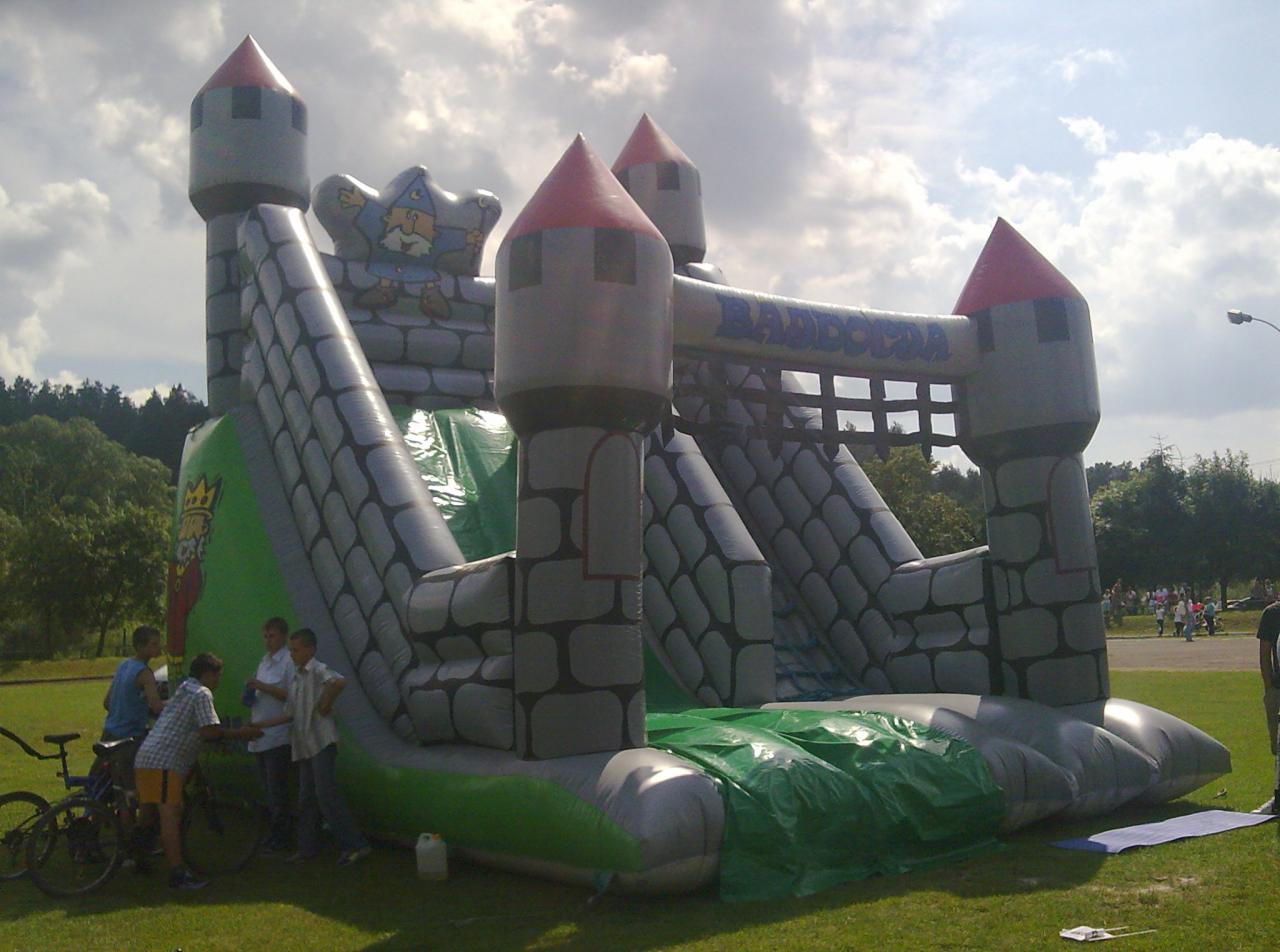 Duża, dmuchana zjeżdżalnia w kształcie zamku z wieżami i napisem 'BADDOCJA', na trawiastym terenie, obok stoi grupa dzieci i rowery, w tle widoczne drzewa i ludzie.