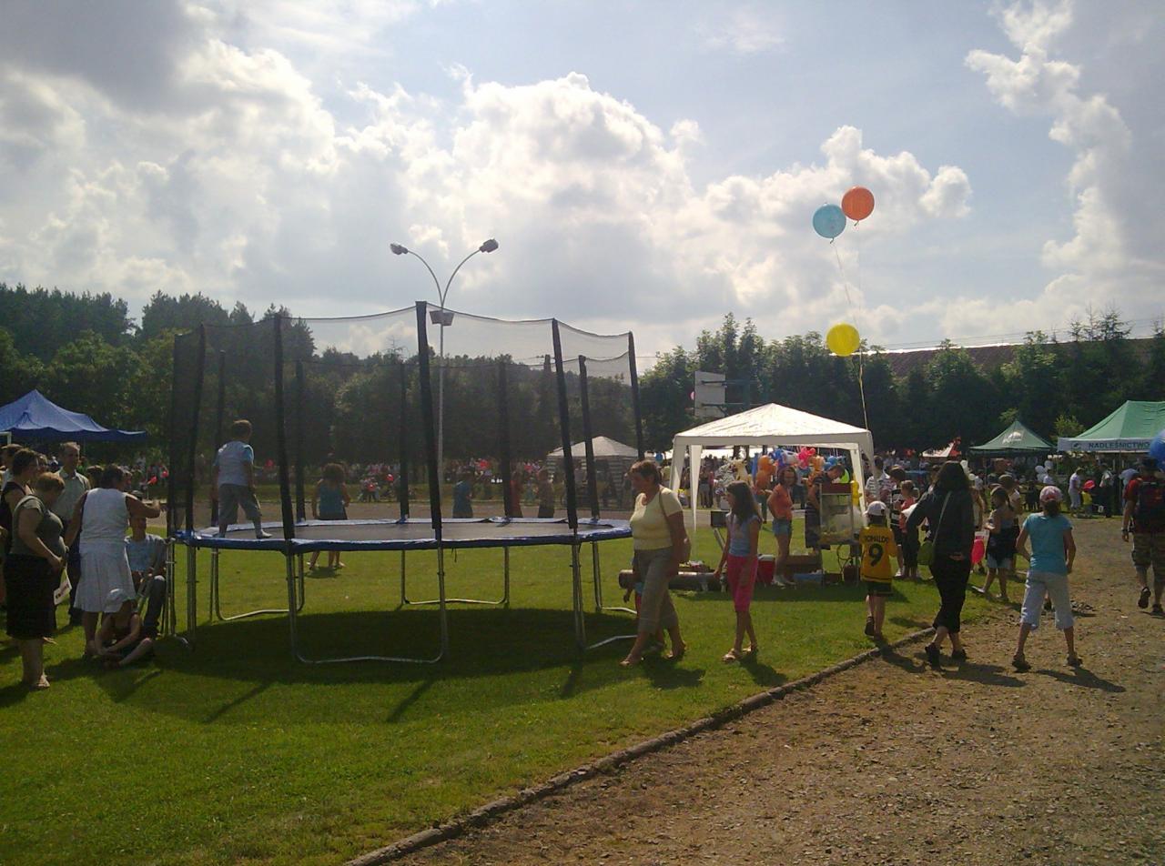 Dzieci skaczą na trampolinie z siatką zabezpieczającą podczas imprezy plenerowej z namiotami i balonami w tle.