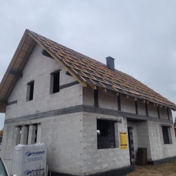 MAR-DOM Usługi Ogólnobudowlane - Surowy stan domu z białych bloczków, widoczna konstrukcja dachu z czarną membraną i drewnianymi łatami, palety bloczków Prefabet przy samochodzie.