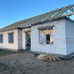 MAR-DOM Usługi Ogólnobudowlane - Budowa domu z białych bloczków, widoczne okna bez szyb, częściowo wykonana więźba dachowa z jasnego drewna, mieszarka do betonu obok budynku.