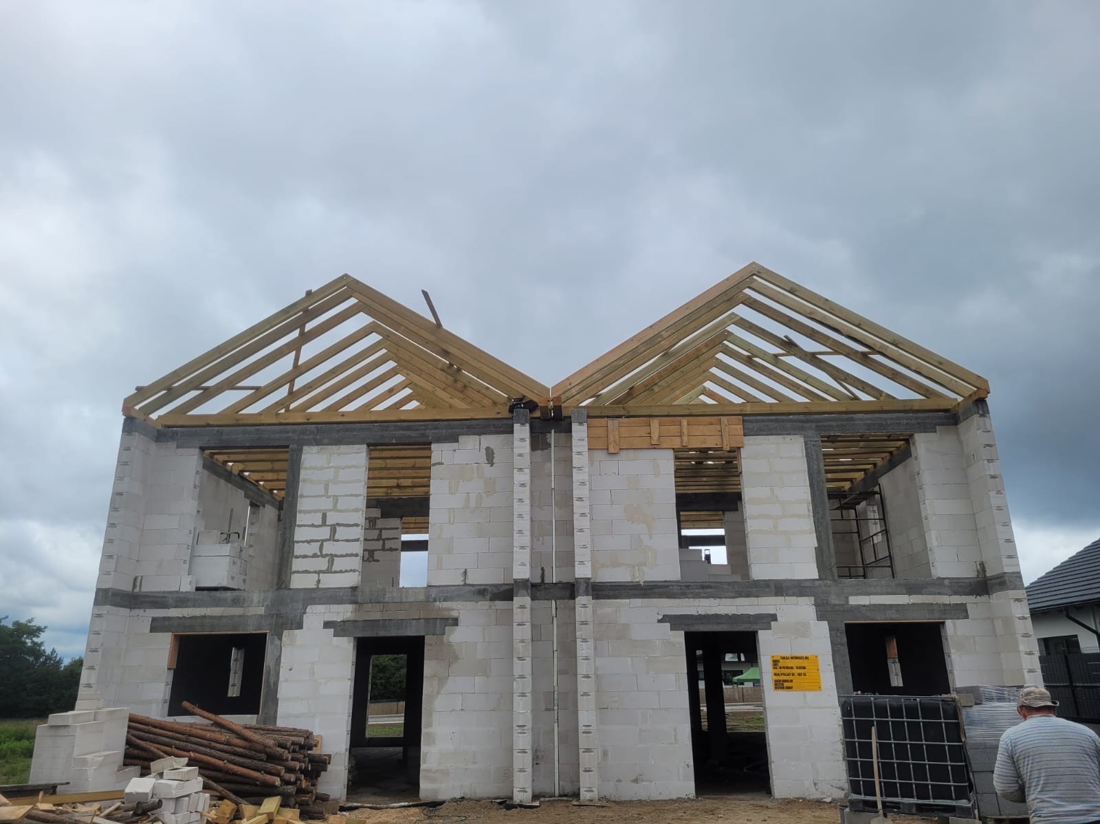 Budowa bliźniaka z białych bloczków z widoczną drewnianą konstrukcją dachu, bez pokrycia, na tle pochmurnego nieba.