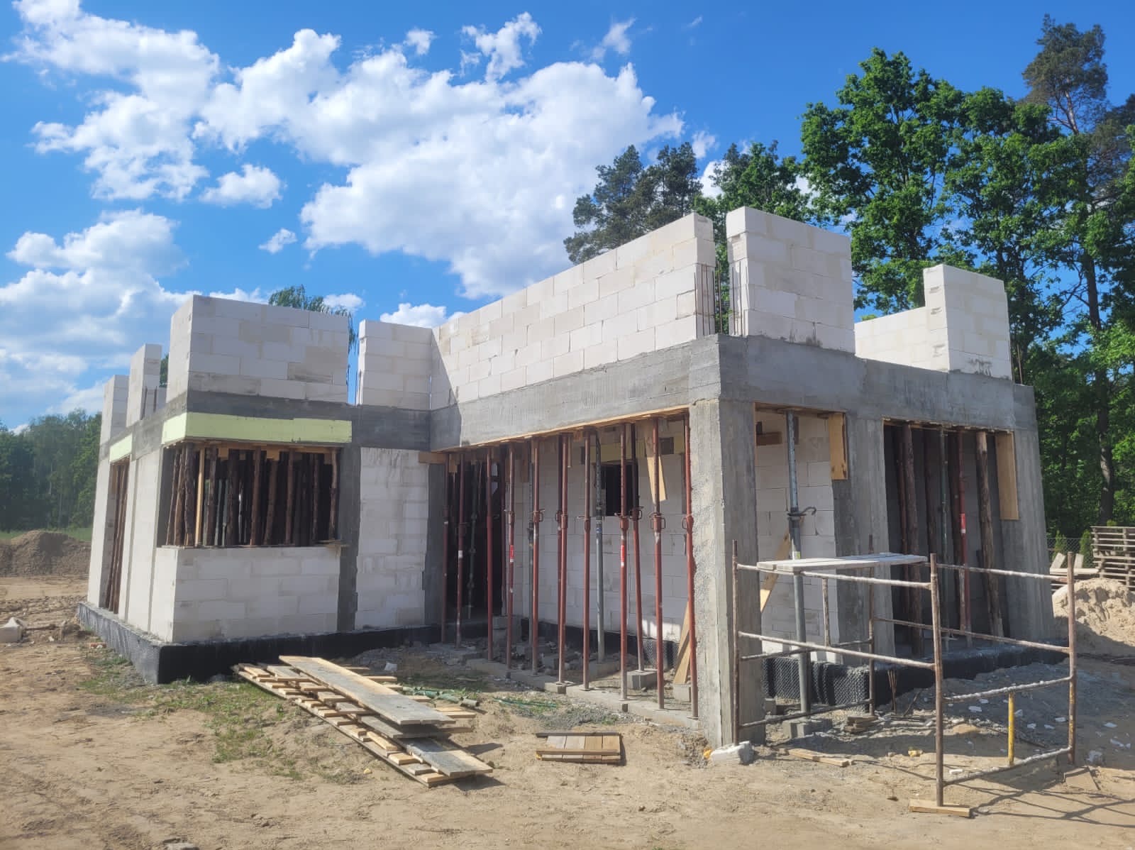 Surowy stan domu z betonu komórkowego, widoczne zbrojenia i podpory szalunkowe, tło błękitne niebo z chmurami.