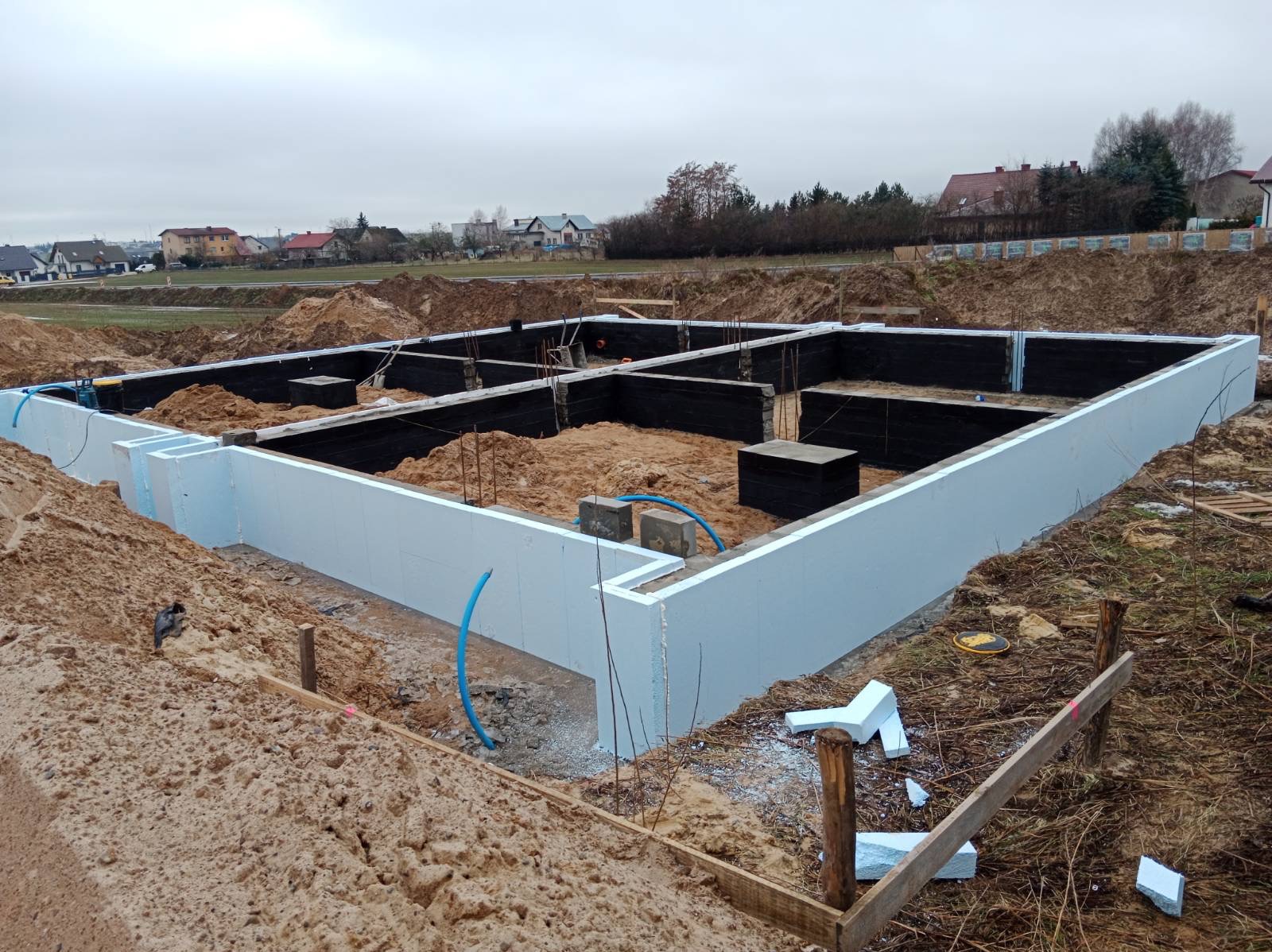 Fundament domu w trakcie budowy: białe ściany szalunkowe z izolacją termiczną, czarna hydroizolacja ścian wewnątrz wykopu, zbrojenie i piasek wewnątrz, teren budowy z ziemią i fragmentami...