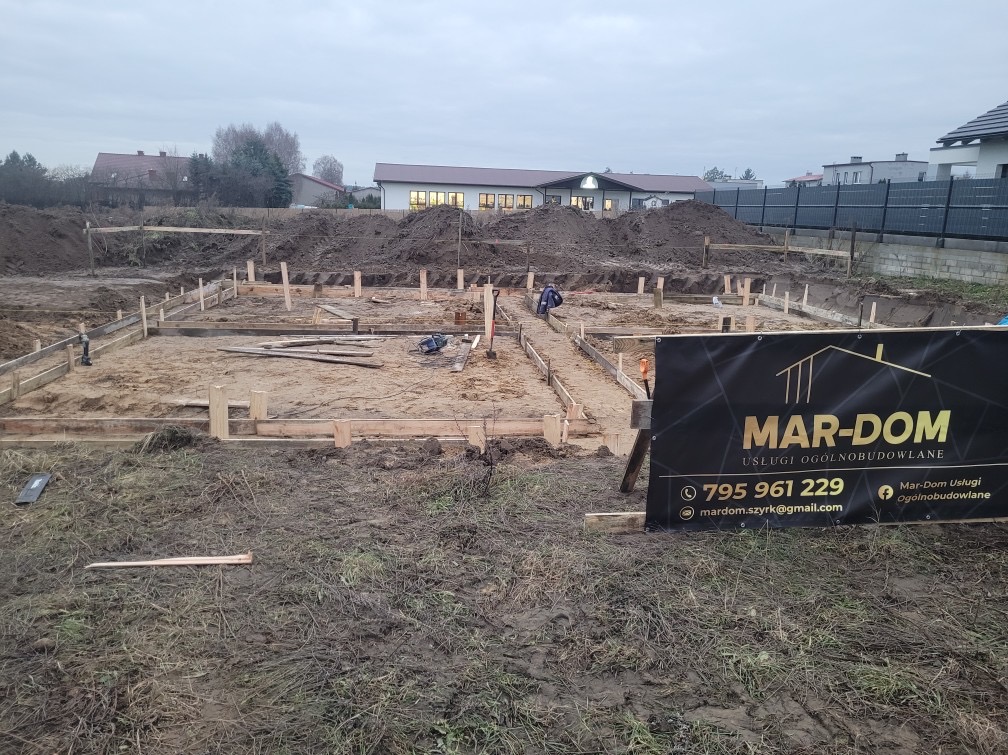 Przygotowanie terenu pod budowę domu jednorodzinnego: wykopane fundamenty z szalunkami, widoczny pracownik i tablica reklamowa firmy MAR-DOM.