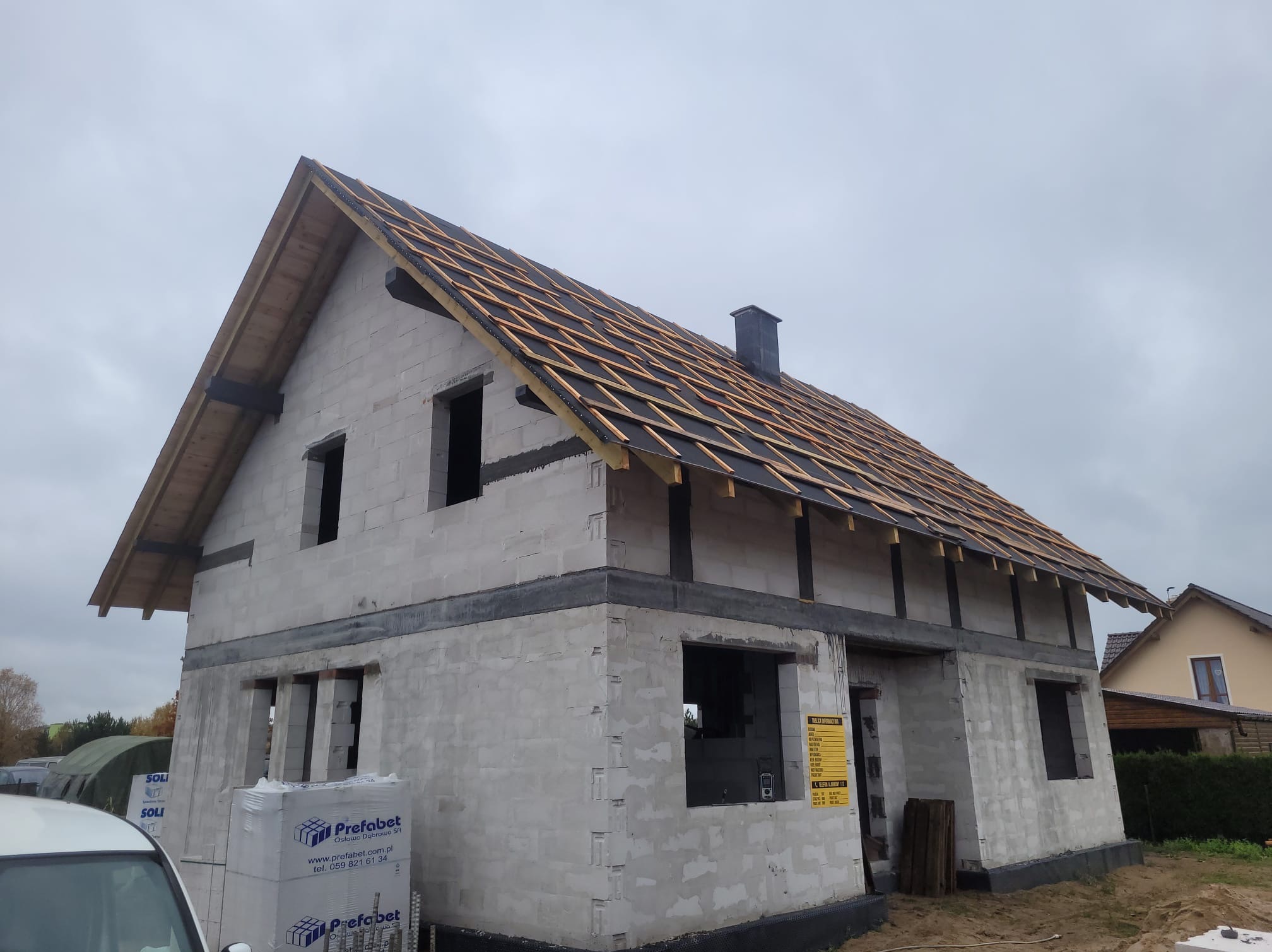 Surowy stan domu z białych bloczków, widoczna konstrukcja dachu z czarną membraną i drewnianymi łatami, palety bloczków Prefabet przy samochodzie.