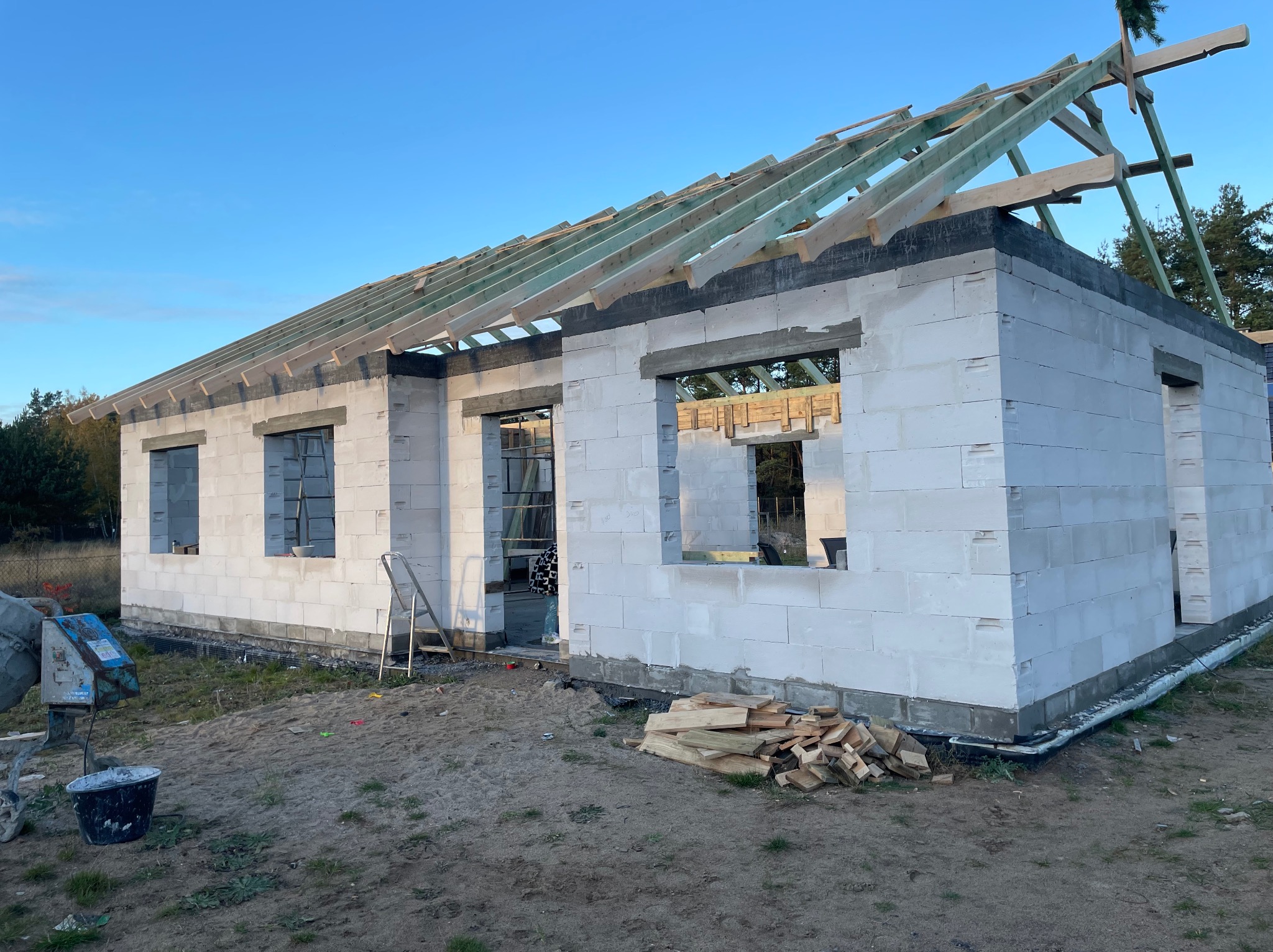 Budowa domu z białych bloczków, widoczne okna bez szyb, częściowo wykonana więźba dachowa z jasnego drewna, mieszarka do betonu obok budynku.