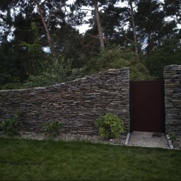 Kamienne Ogrody - Kamienny mur z brązową furtką wkomponowany w zieleń ogrodu, widoczne małe krzewy i trawnik.