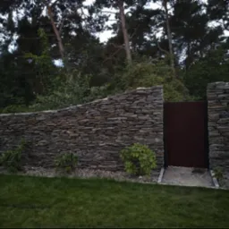 Kamienny mur z brązową furtką wkomponowany w zieleń ogrodu, widoczne małe krzewy i trawnik.