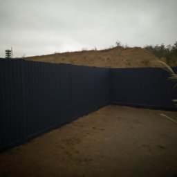 Kamienne Ogrody - Ciemny, panelowy płot okalający teren z widocznym pagórkiem i wieżą obserwacyjną w tle, częściowo zasłonięty trawą pampasową.