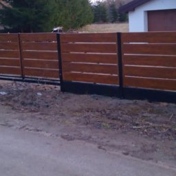Kamienne Ogrody - Nowoczesne ogrodzenie z poziomych desek w kolorze drewna, z czarnymi słupkami i podmurówką, widoczne z perspektywy ulicy.