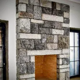 Kominek obłożony nieregularną kostką granitową w odcieniach szarości i beżu, z widocznym wnętrzem paleniska wyłożonym cegłą, w tle okna z czarnymi ramami.