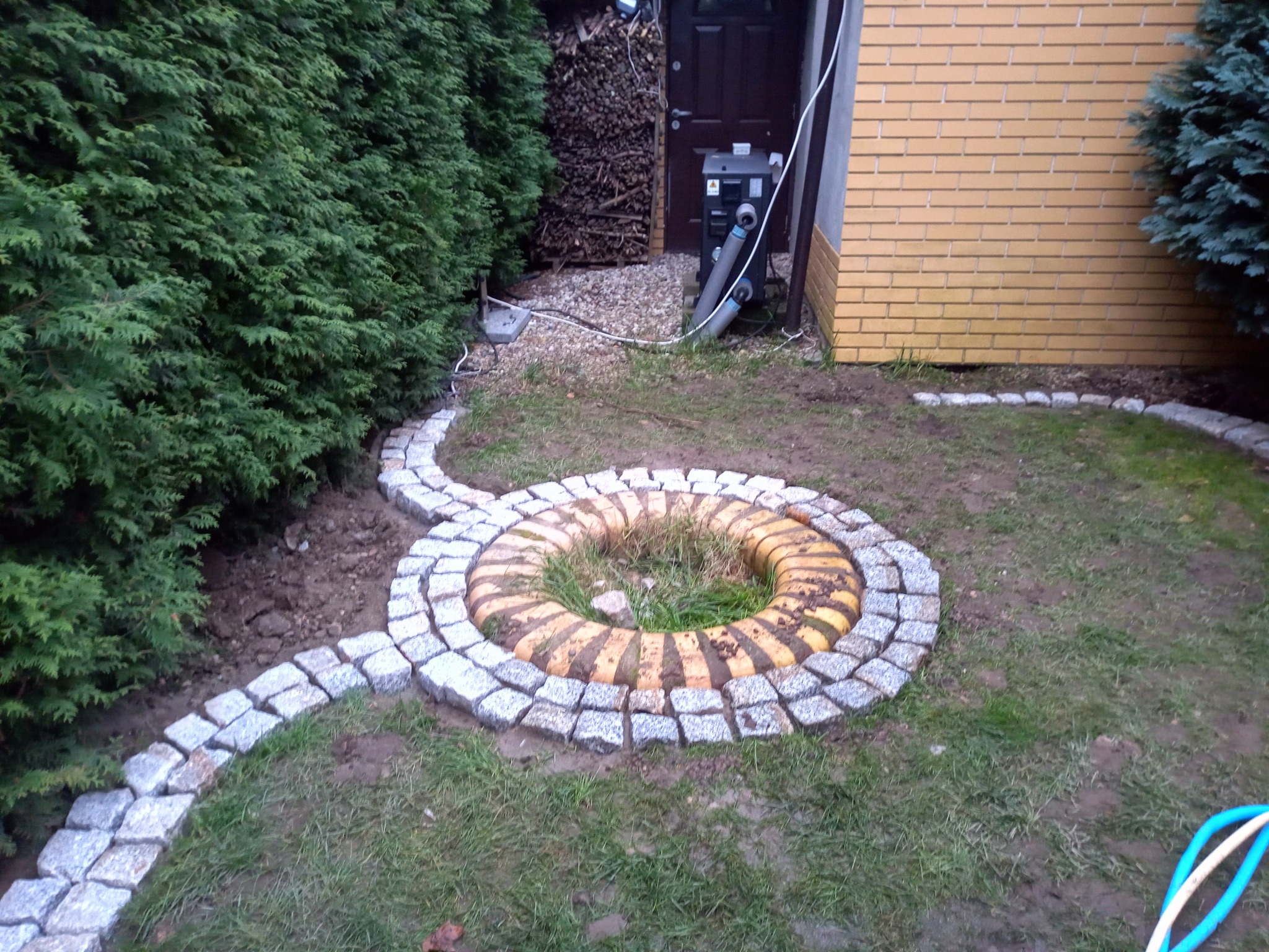 Ścieżka z kostki granitowej prowadząca do okrągłego klombu z obrzeżem z kostki, tło: zielony żywopłot i ściana budynku.