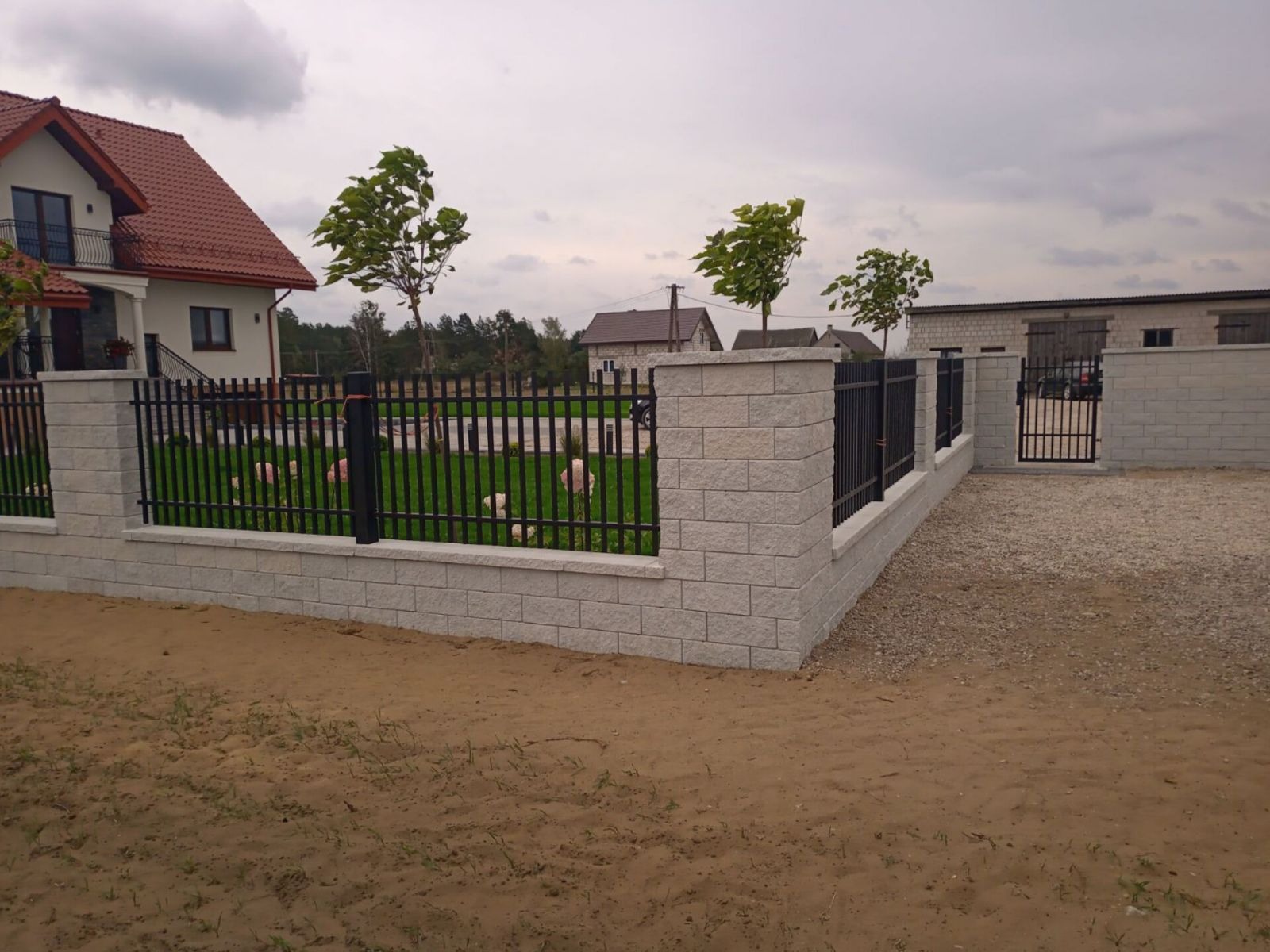 Nowoczesne ogrodzenie z szarego kamienia łupanego i czarnych metalowych przęseł, widok z zewnątrz posesji na tle domu z czerwonym dachem i zielonego trawnika.