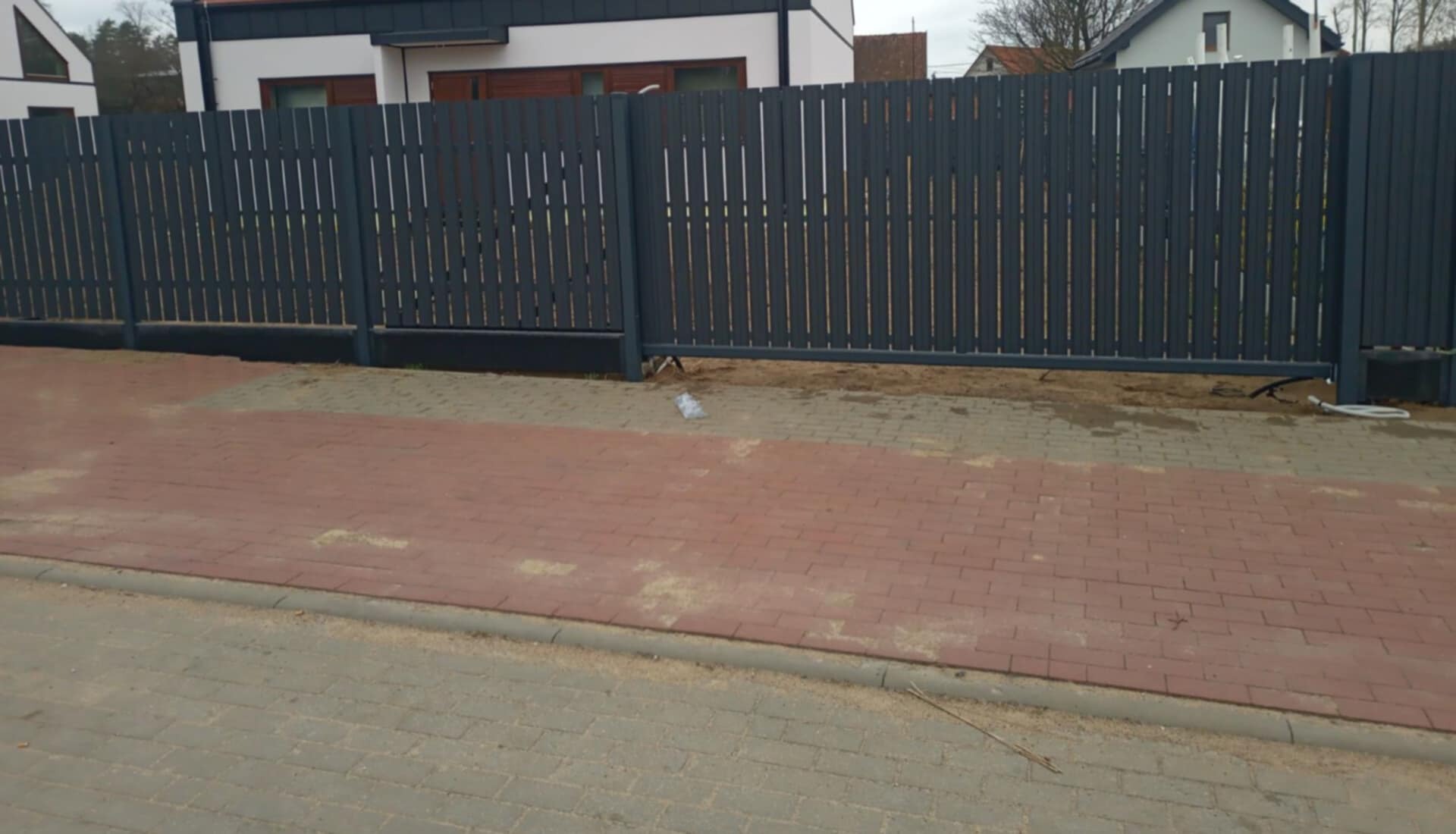 Nowoczesne, szare ogrodzenie panelowe z przesuwną bramą, widoczne od strony posesji, z fragmentem kostki brukowej i domu w tle.