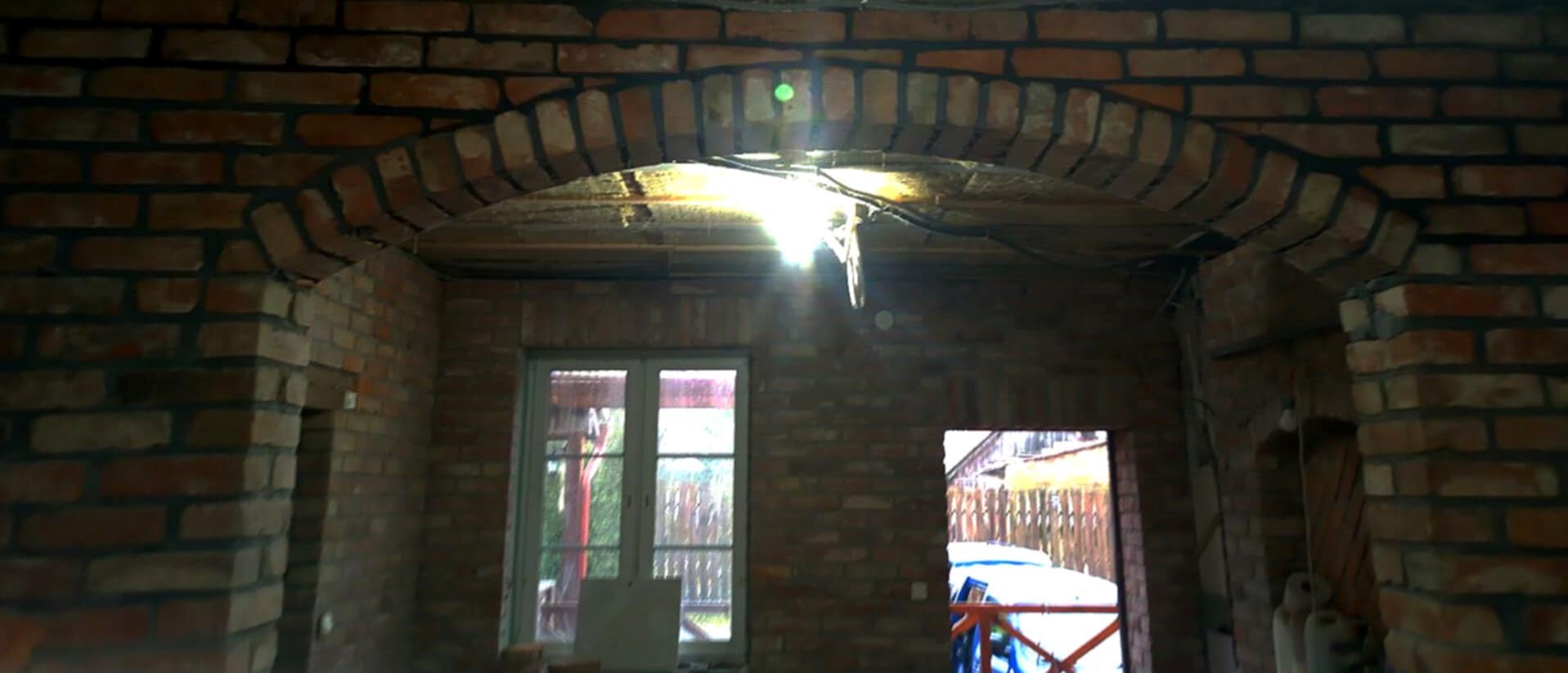 Wnętrze budynku w trakcie budowy z widocznym murowanym łukiem nad oknem i drzwiami, cegły w odcieniach czerwieni i brązu, widoczne przewody elektryczne i surowe wykończenie.