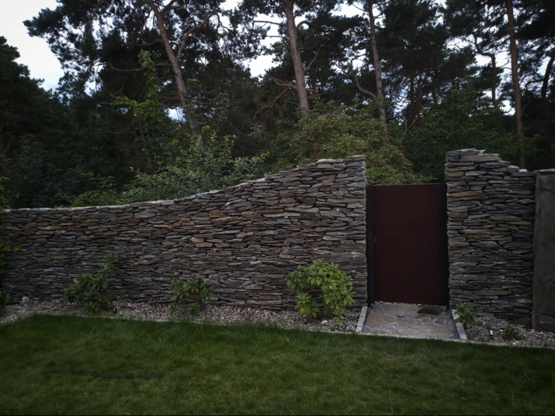 Kamienny mur z brązową furtką wkomponowany w zieleń ogrodu, widoczne małe krzewy i trawnik.