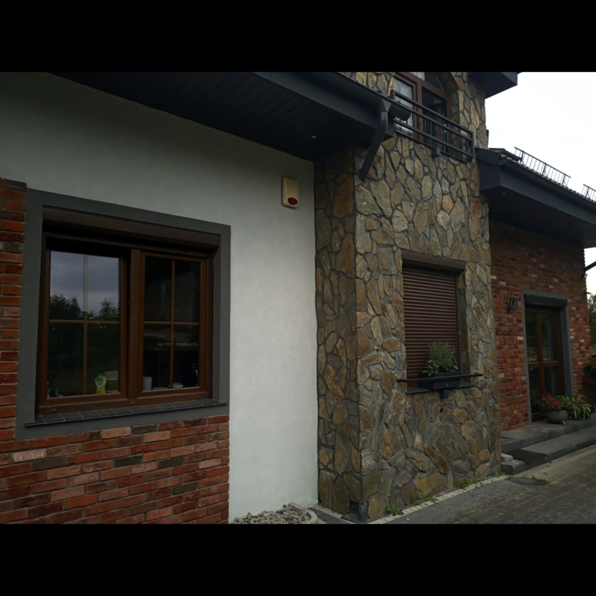 Fragment elewacji domu z elementami z kamienia, cegły klinkierowej i jasnego tynku, okna z drewnianą stolarką i roletami zewnętrznymi, doniczka z kwiatami pod oknem.