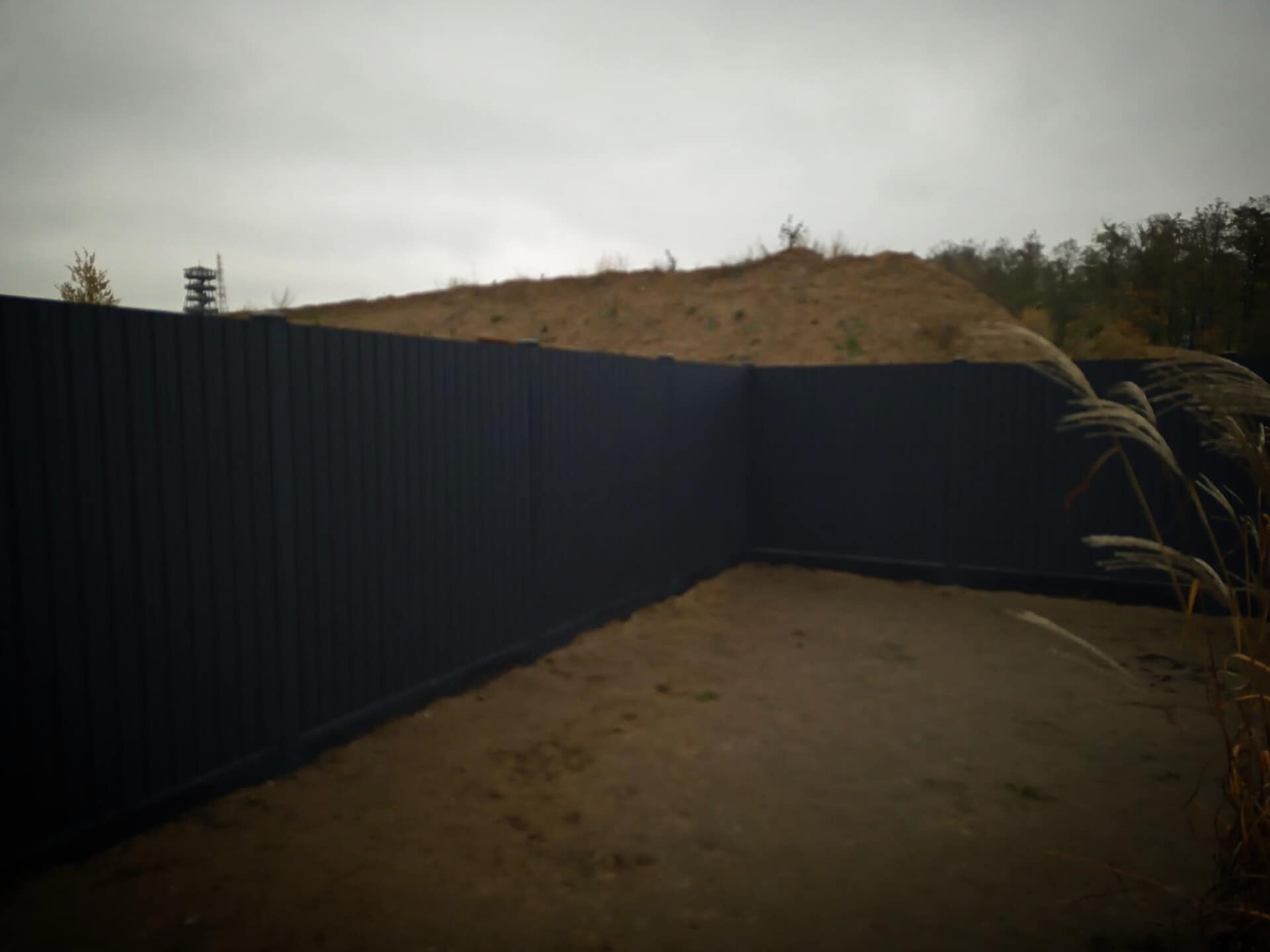 Ciemny, panelowy płot okalający teren z widocznym pagórkiem i wieżą obserwacyjną w tle, częściowo zasłonięty trawą pampasową.