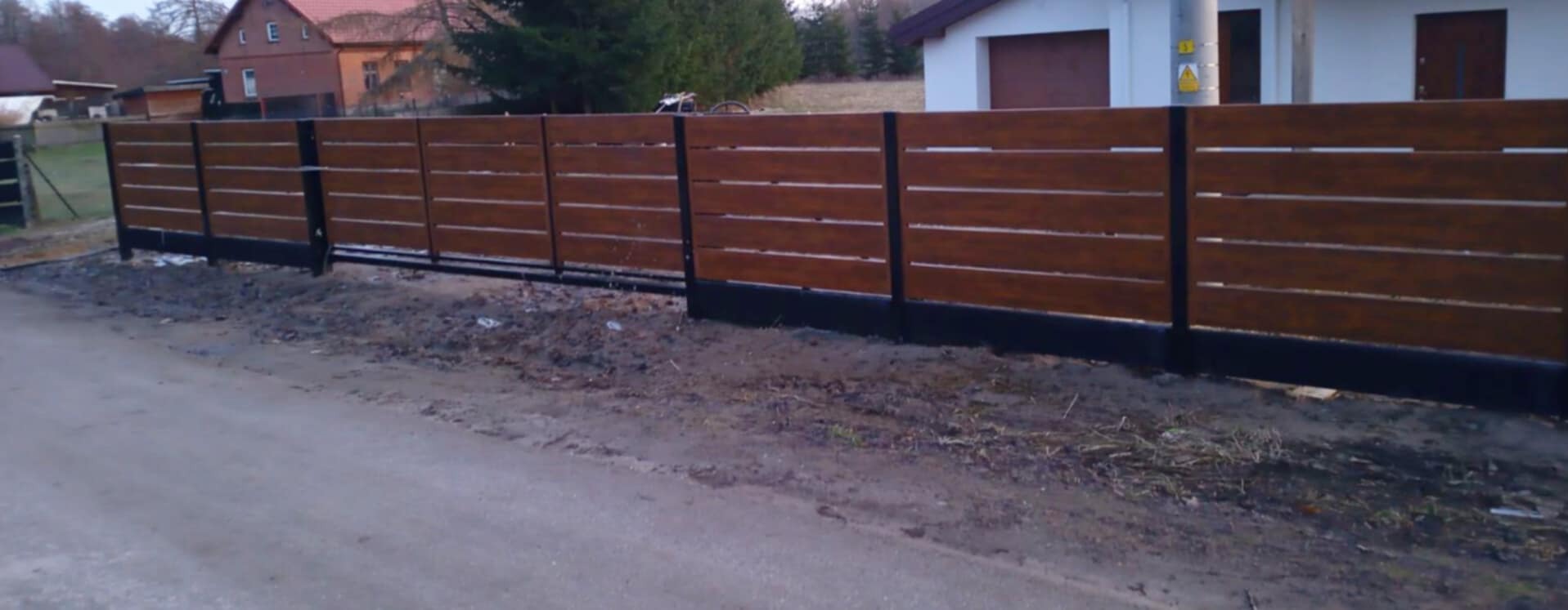 Nowoczesne ogrodzenie z poziomych desek w kolorze drewna, z czarnymi słupkami i podmurówką, widoczne z perspektywy ulicy.