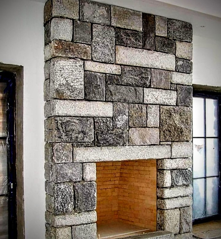 Kominek obłożony nieregularną kostką granitową w odcieniach szarości i beżu, z widocznym wnętrzem paleniska wyłożonym cegłą, w tle okna z czarnymi ramami.