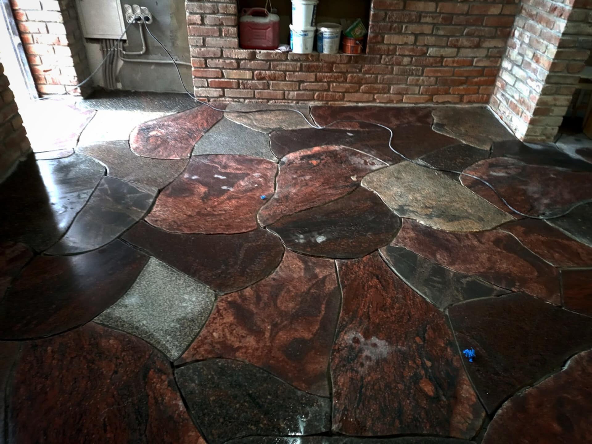 Podłoga wyłożona nieregularnymi płytami z kamienia w odcieniach brązu i szarości, z widoczną instalacją elektryczną i fragmentem ściany z czerwonej cegły w tle.