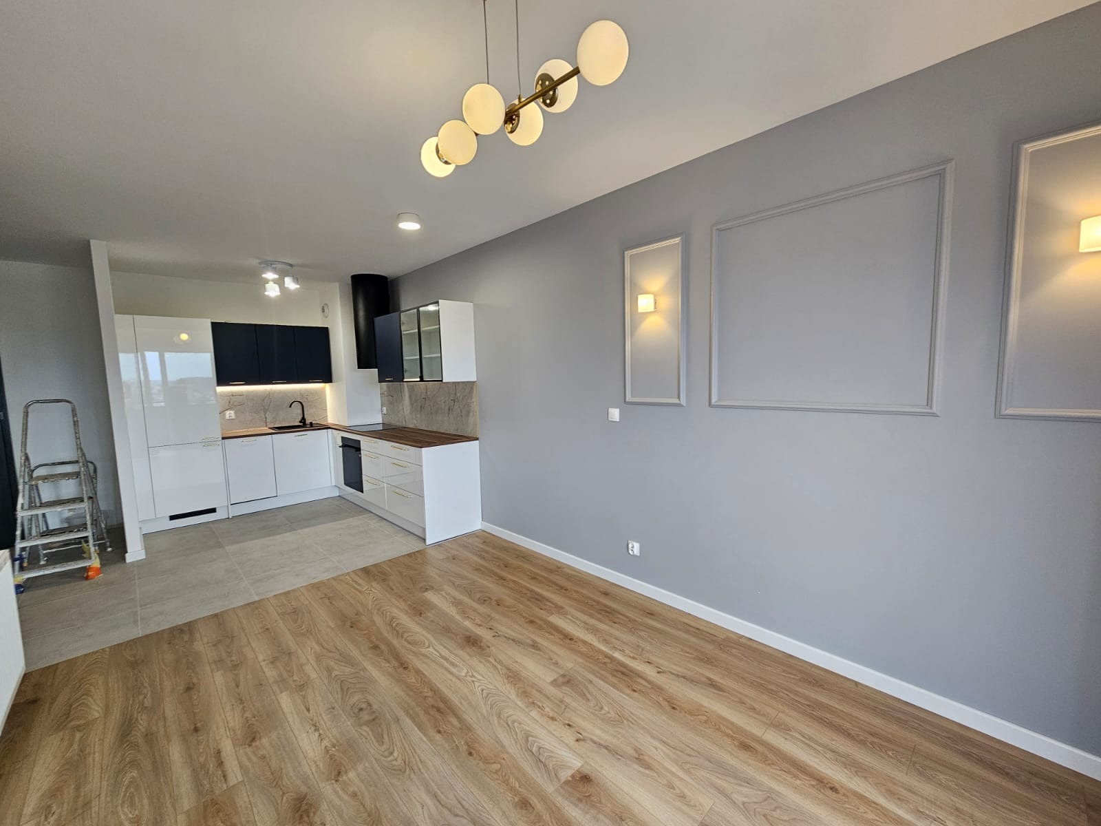 Wykończone wnętrze mieszkania z drewnianą podłogą, szarą ścianą z dekoracyjnymi ramkami, nowoczesną kuchnią z białymi i granatowymi szafkami oraz złotymi uchwytami.